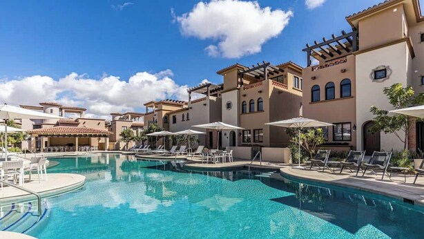 Un área de piscina con sillones de descanso y sombrillas, rodeada de edificios de estilo mediterráneo.