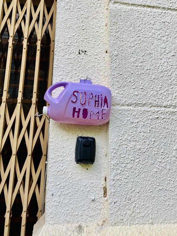A purple mailbox with "SOPHIA HOME" written on it, attached to a textured wall next to a window with a metal grille.