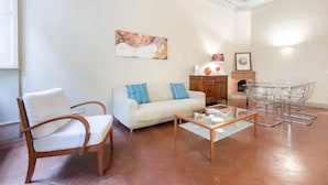 A living room with a white sofa, a wooden chair, a glass coffee table, and a dining area with clear chairs. - Reinassance Beauty - elegant apartment in the heart of Florence (Firenze)