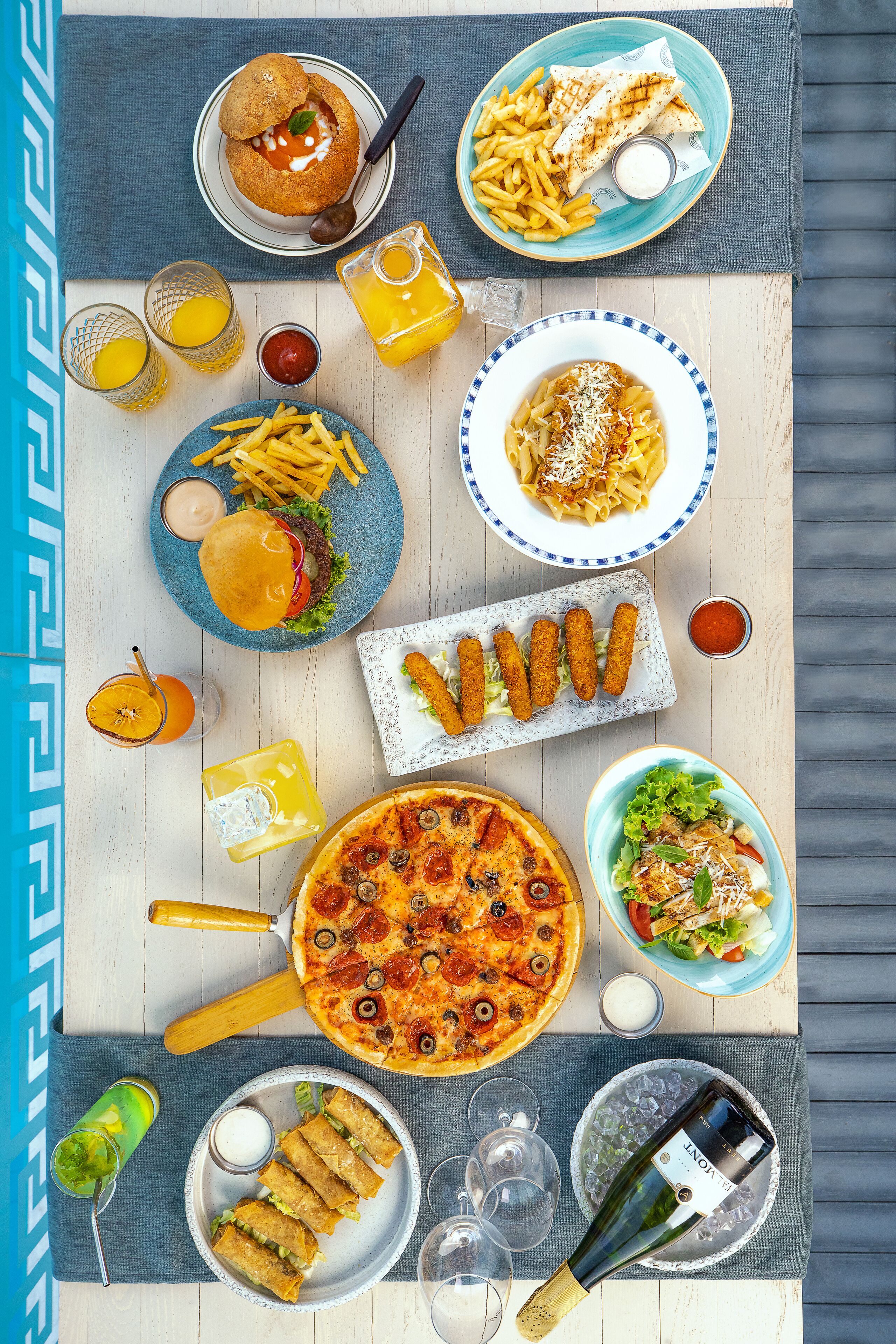 A table with various dishes including pizza, pasta, and fried items, along with drinks and a bottle of wine.
