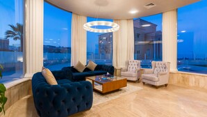 A modern living room with a blue sofa, a wooden coffee table, and striped armchairs. Large windows offer a view of a building and palm trees. - Selena Bay Resort (Hurghada)