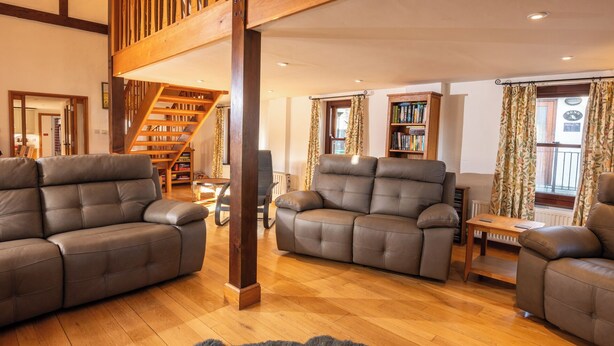 A spacious living room with a wooden staircase, leather sofas, a bookshelf, and a view of the outdoors. - Keiras Cottage - Three Bedroom House, Sleeps 6 (Ambleside)