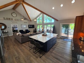 A modern kitchen with a central island, a dining area with bar stools, a living room with a sofa, and a large window offering a view of the outdoors.