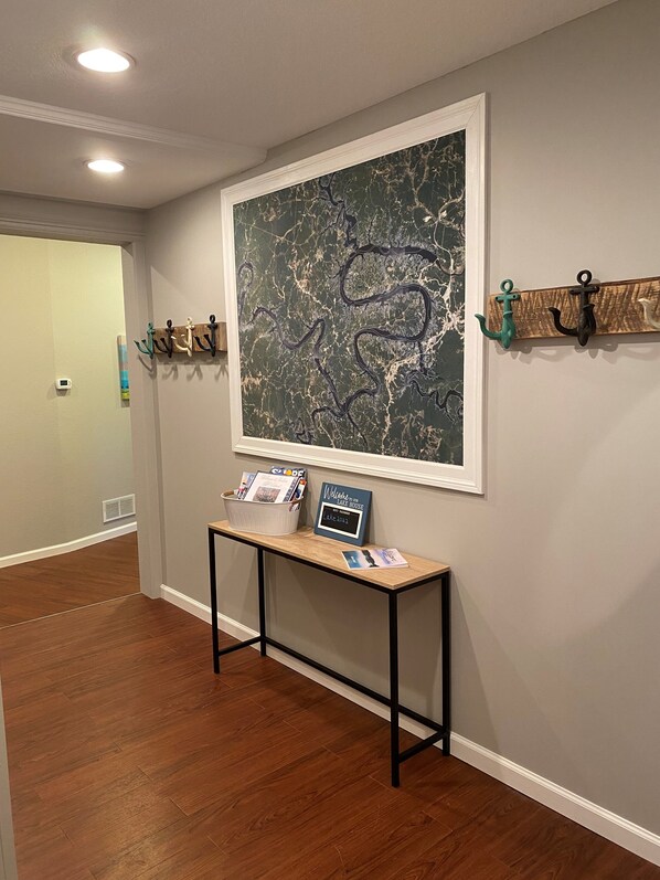 A hallway with a wooden floor, a framed abstract painting, a small table with a welcome sign, and two coat hooks with decorative items. - Large, private, multifamily lake retreat. (Sunrise Beach)