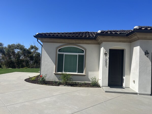 A single-story house with a large arched window, a dark door, and a small garden area with plants and a concrete driveway. - Brand New  Carlsbad Suite Near  La Costa  Resort (Carlsbad)