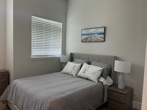 A bedroom with a bed, two bedside tables, a window with blinds, a painting on the wall, and a nightstand with a lamp. - Brand New  Carlsbad Suite Near  La Costa  Resort (Carlsbad)
