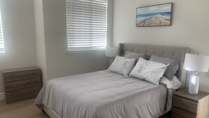 Une chambre à coucher avec un lit, des tables de chevet, une commode, un tableau au mur et une fenêtre avec des stores.