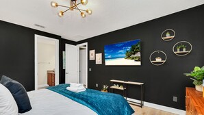 Un dormitorio con cama, cubrecama azul, televisor montado en la pared y una pequeña estantería con plantas.