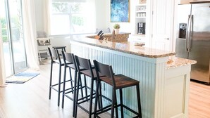 Una cocina moderna con una isla central, taburetes de bar, iluminación colgante y un refrigerador de acero inoxidable.