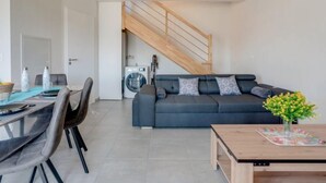 A modern living room with a dark grey sofa, a wooden coffee table, and a dining area with a glass table and chairs. A washing machine is visible in the background, and a wooden staircase leads to another level. - Soleil Couchant B03 by Interhome (Mimizan)