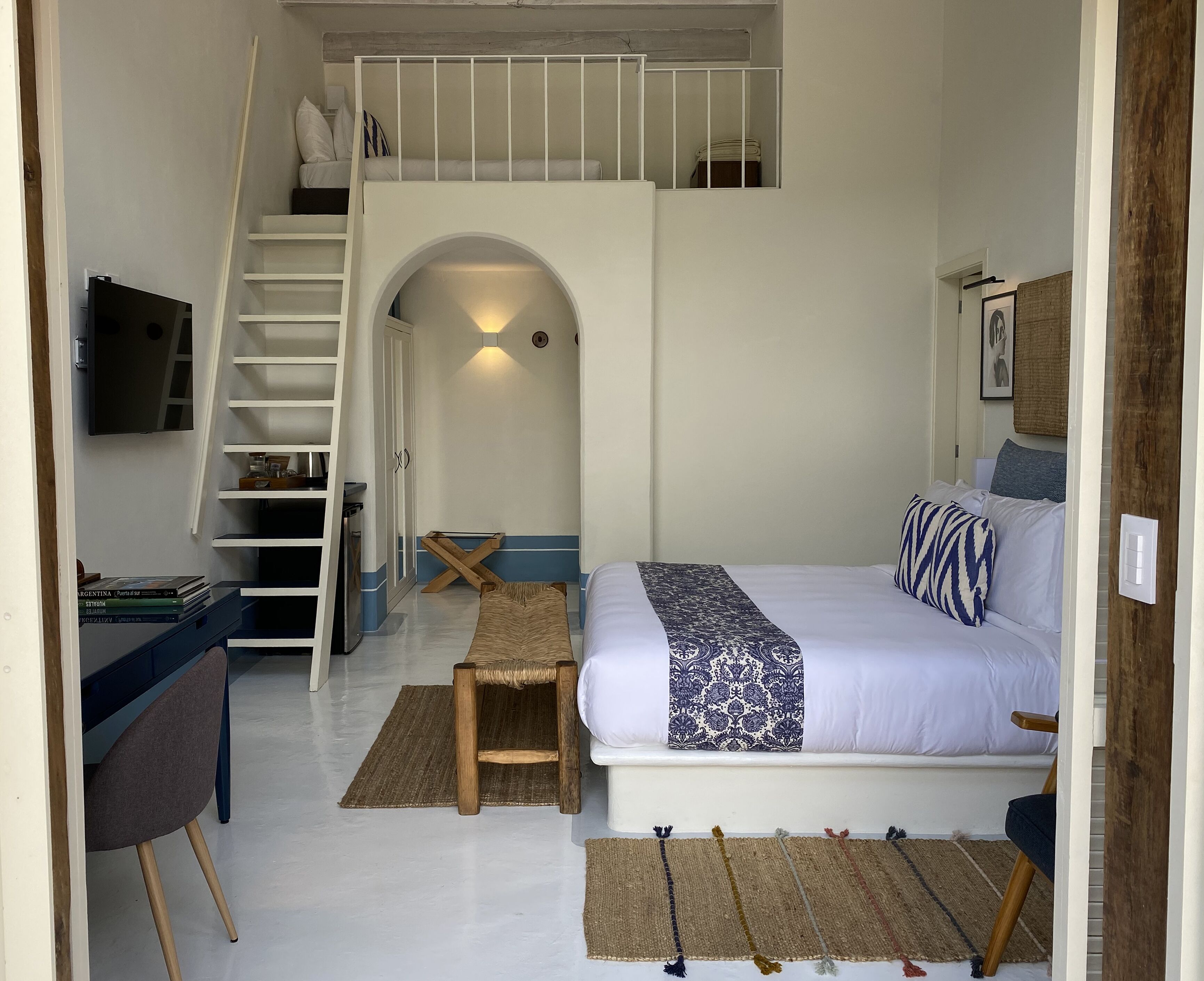 A bedroom with a bed, a desk, a chair, a bench, a staircase, and a small table.