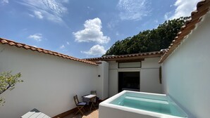 Un pequeño espacio de piscina al aire libre con una silla y una mesa, rodeado de paredes blancas y un techo de tejas.