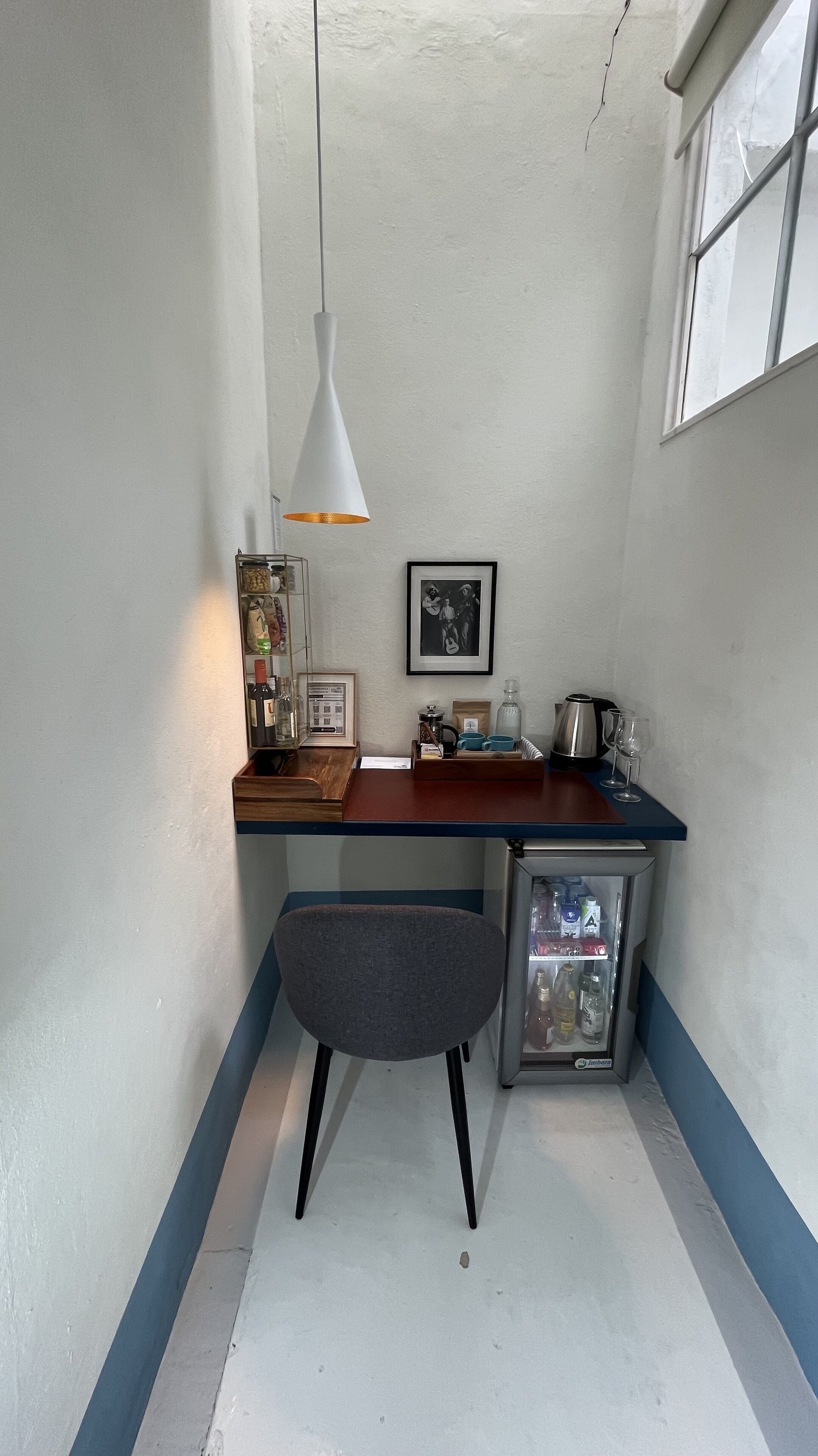 Una cocina compacta con encimera de madera, un refrigerador pequeño, una silla gris, una lámpara colgante y una ventana.