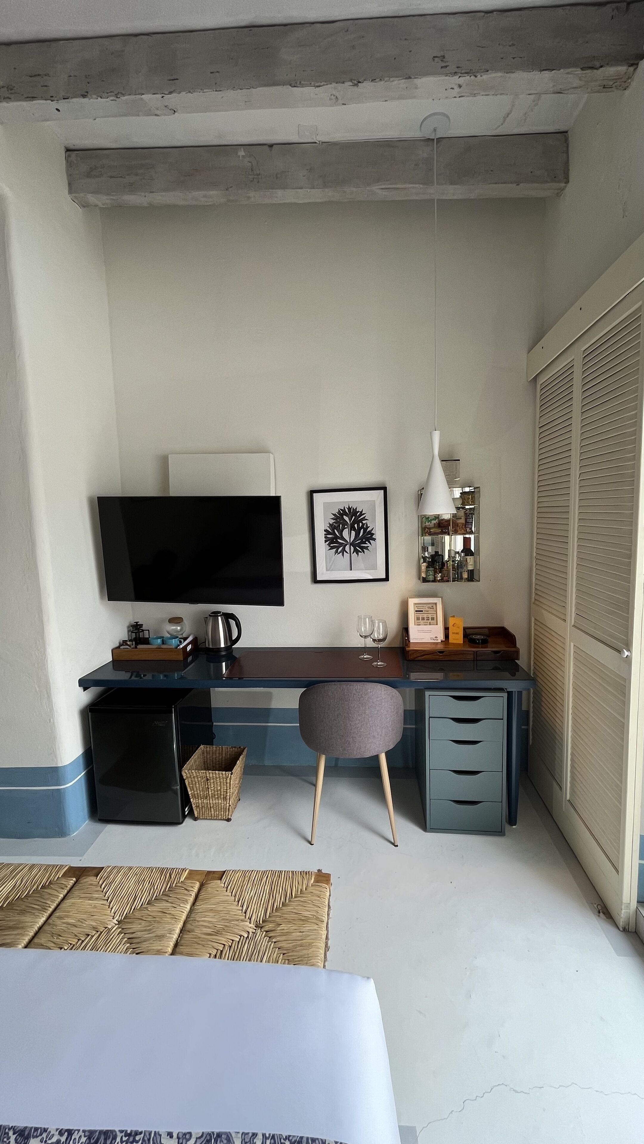A modern hotel room with a flat-screen TV, a framed artwork, a desk with a lamp, a minibar, and a large window with blinds.