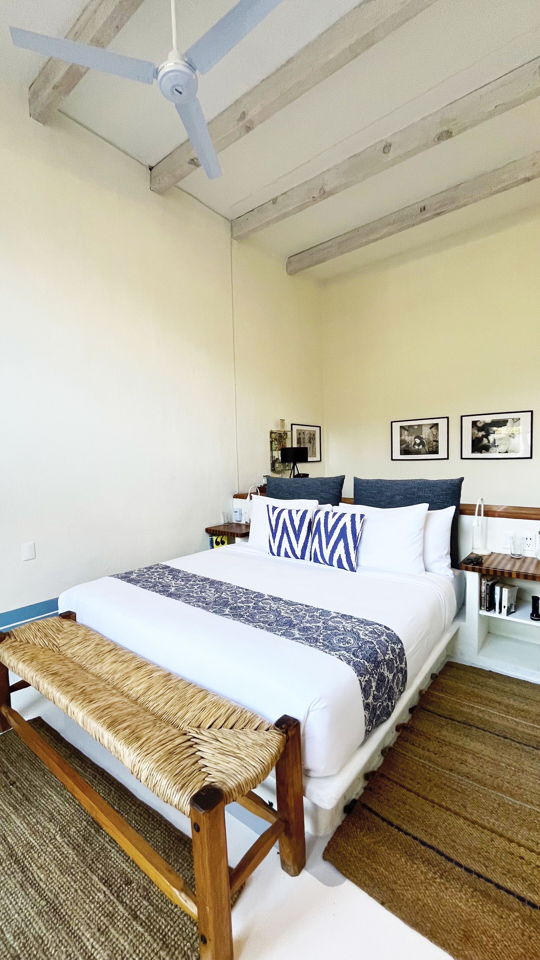 A neatly arranged bedroom with a bed, a bench, a ceiling fan, and framed pictures on the wall.