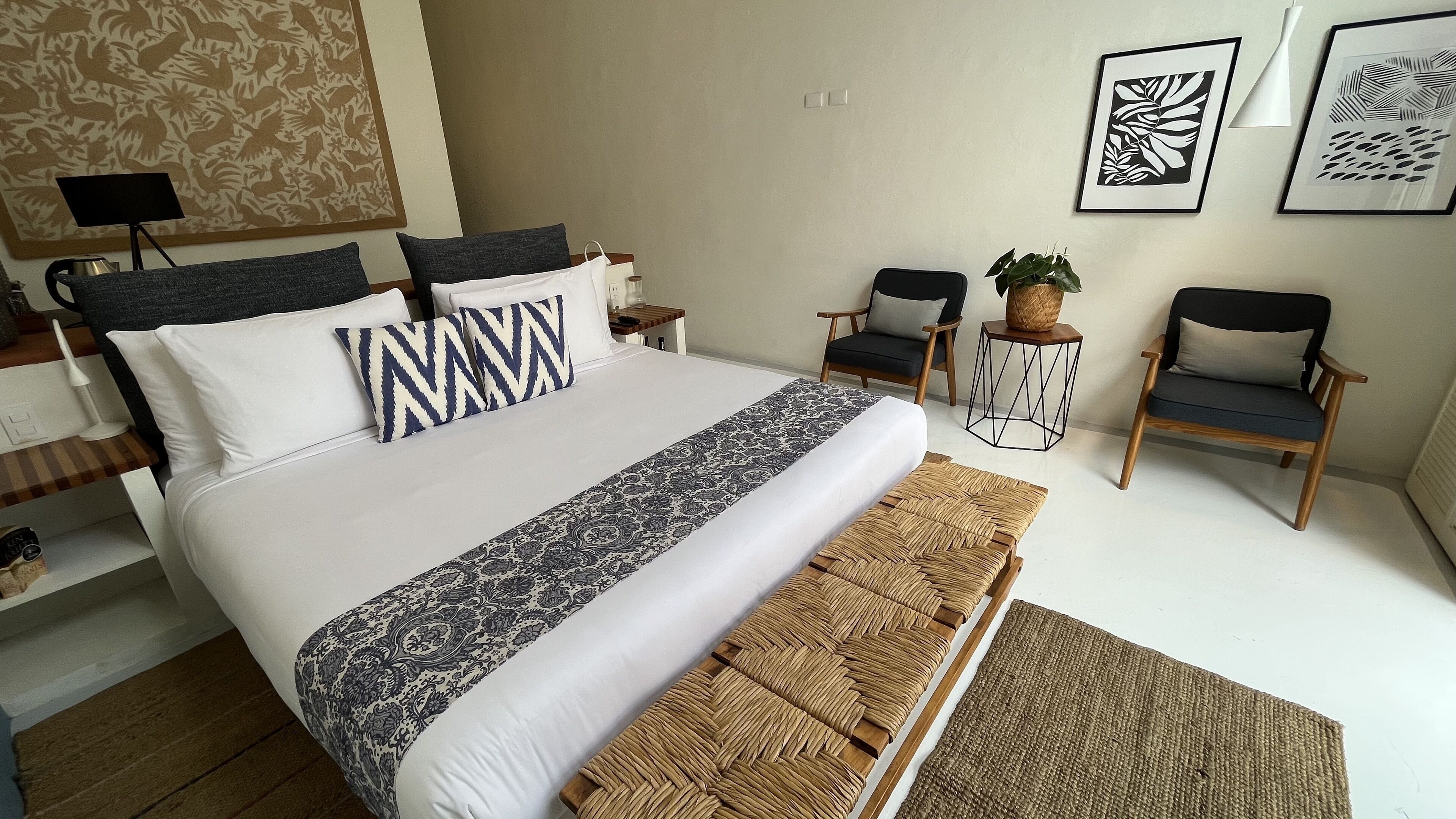 A neatly arranged bedroom with a large bed, two chairs, a small table, and a potted plant.