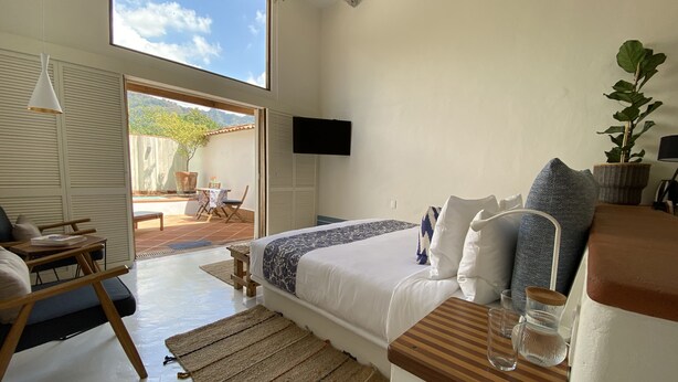 Un dormitorio con cama, mesita de noche y televisor en la pared. Se ve una ventana con vistas al exterior.