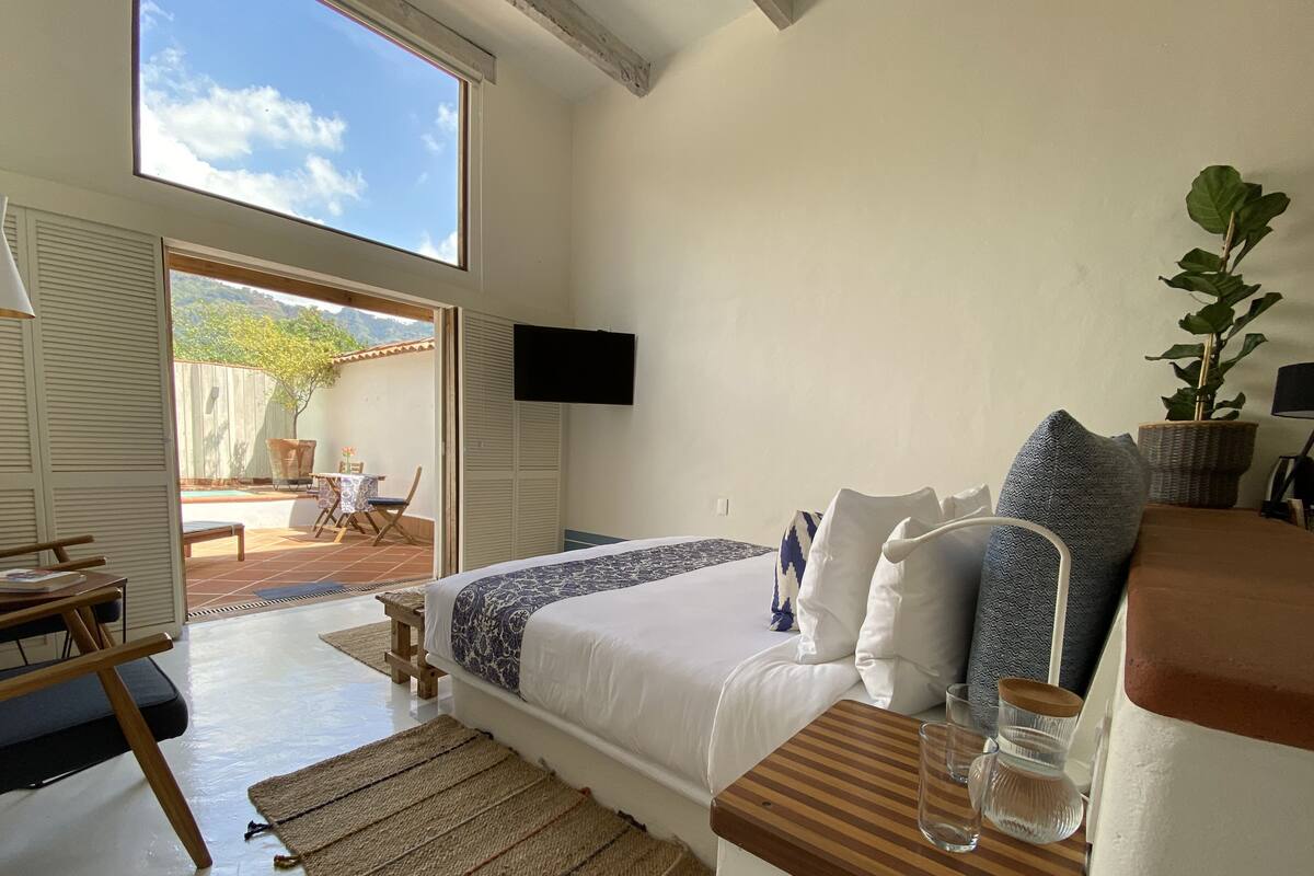 A bedroom with a bed, bedside table, and a TV on the wall. There is a window with a view of the outdoors.