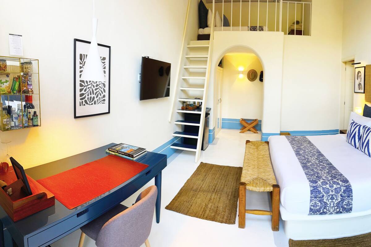 A compact hotel room with a bed, a desk, a chair, a small table, a bookshelf, and a staircase leading to an upper level.