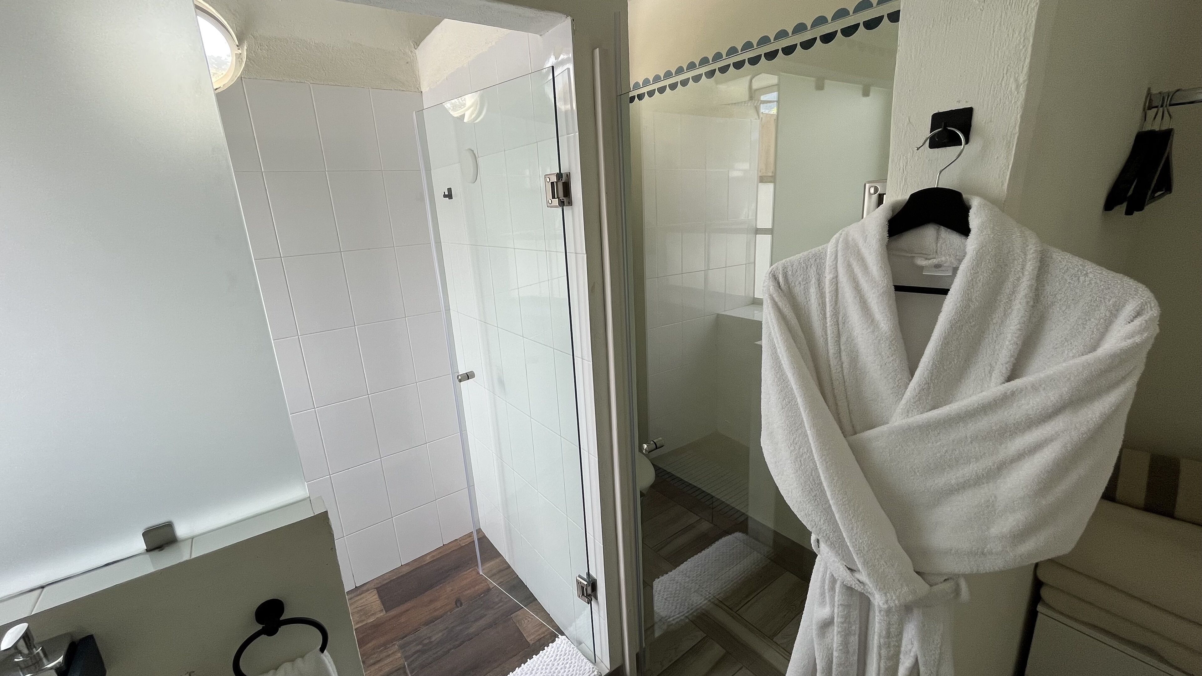 A bathroom with a glass shower door, a white bathrobe hanging on a rack, and a wooden floor.