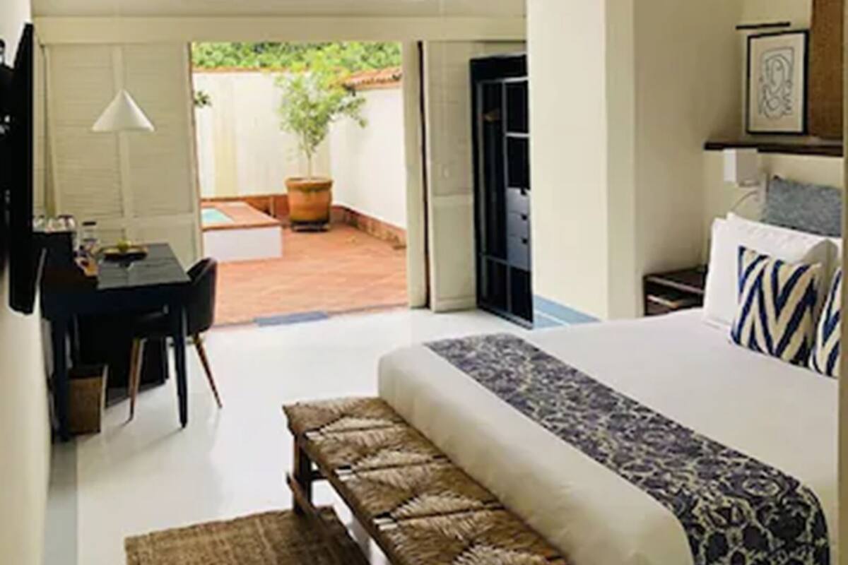 A bedroom with a bed, a bench, a desk, and a window overlooking a garden.