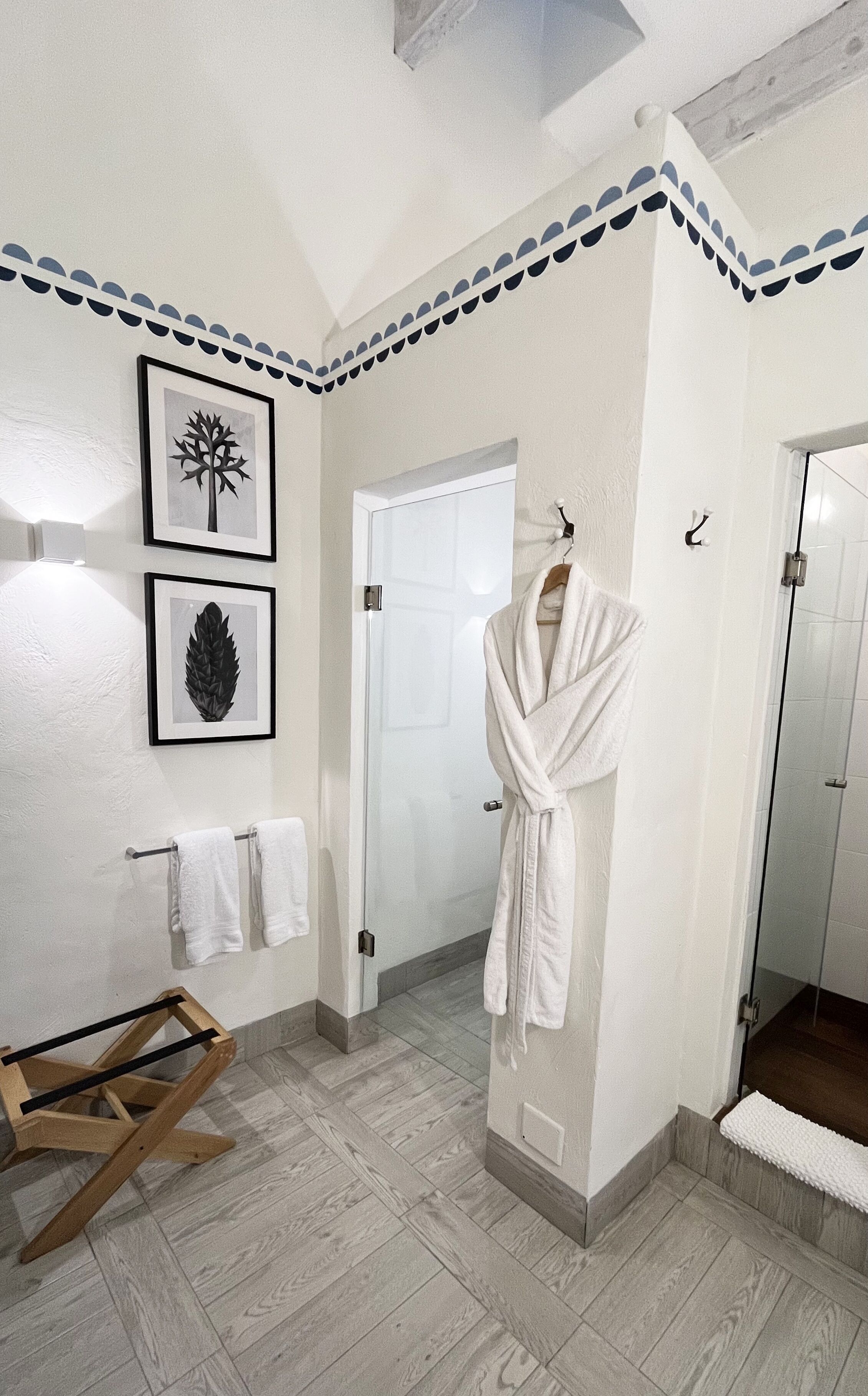 A bathroom with a wooden floor, white walls, a glass shower, and a robe hanging on a hook. There are two framed pictures on the wall.