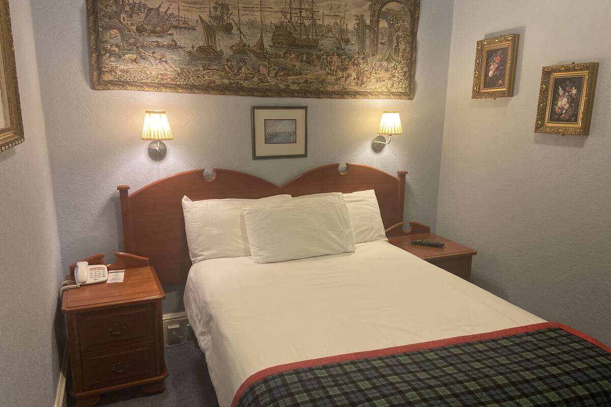 A hotel room with a wooden bed, two bedside lamps, a telephone, a small table, and a painting on the wall.