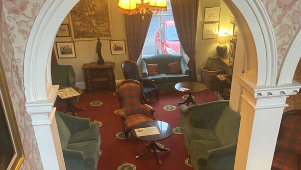 A grand staircase with ornate railings leading to a room with a red carpet, multiple armchairs, a sofa, and a chandelier. - Lovat Arms Hotel (Beauly)