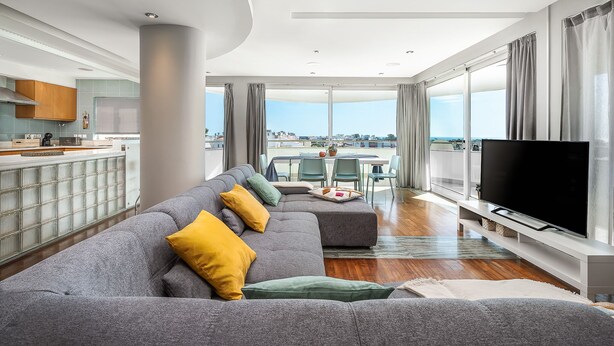 Een moderne woonkamer met een grijze hoekbank, een flatscreen televisie en een eethoek met een tafel en stoelen.