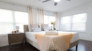 Un dormitorio con cama, mesitas de noche, ventilador de techo y ventanas grandes con cortinas.