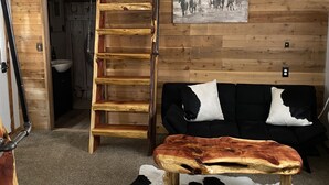 A rustic cabin interior with a loft bed, a wooden ladder, a black and white sofa, a wooden bench, and a cowhide rug. - Deadwood acres cabin #3 (Eminence)