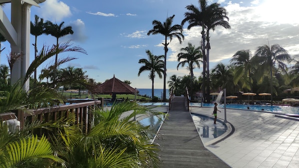 Zona de piscina con palmeras, una terraza de madera y vistas al océano.
