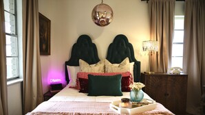 Un lit bien fait, agrémenté d’un jeté rose, une table de chevet avec une lampe et une commode sur laquelle est posé un vase de fleurs.