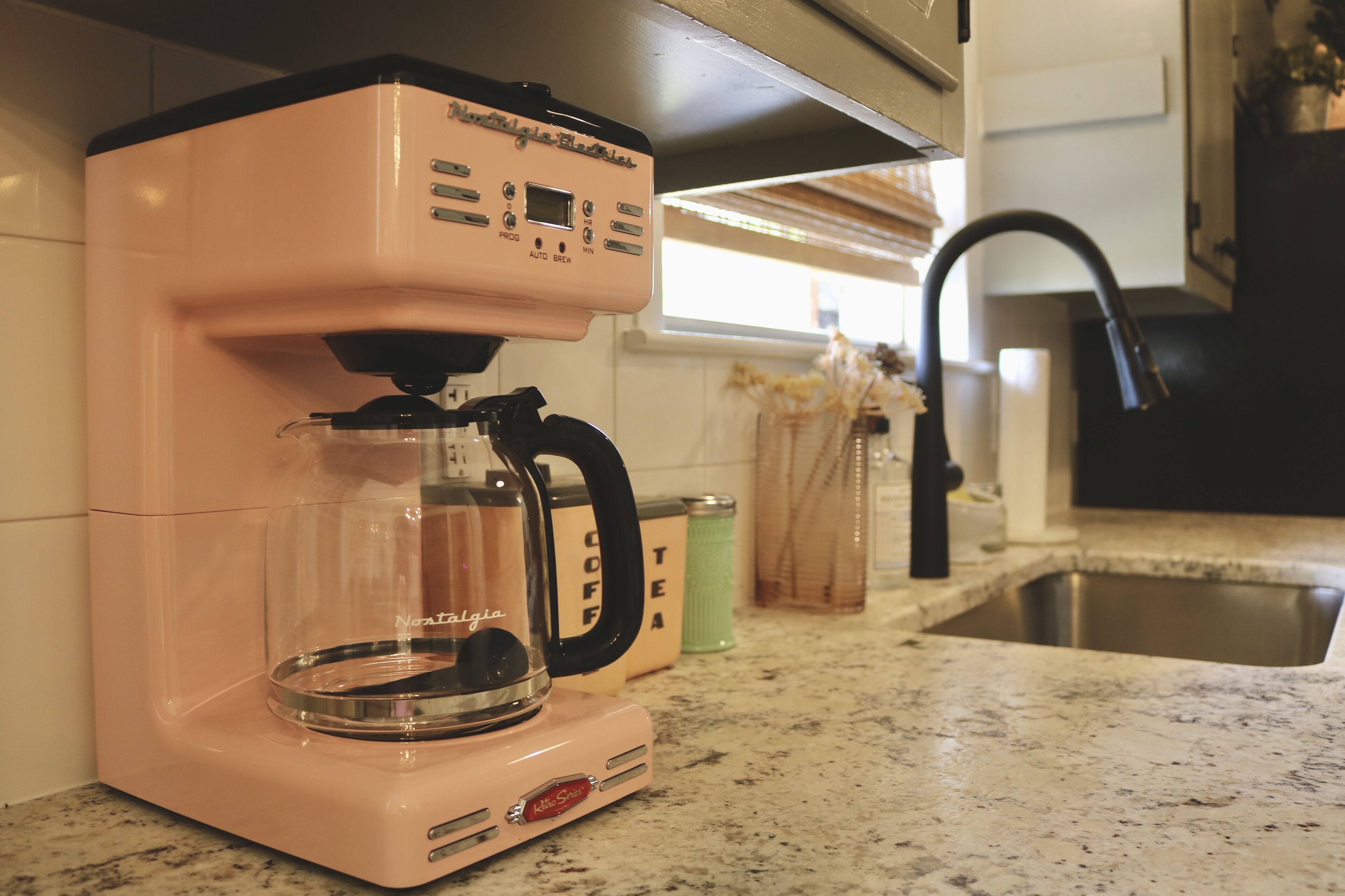 A pink coffee maker on a kitchen counter with a stainless steel sink and a window in the background.