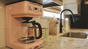 A pink coffee maker on a kitchen counter with a stainless steel sink and a window in the background.