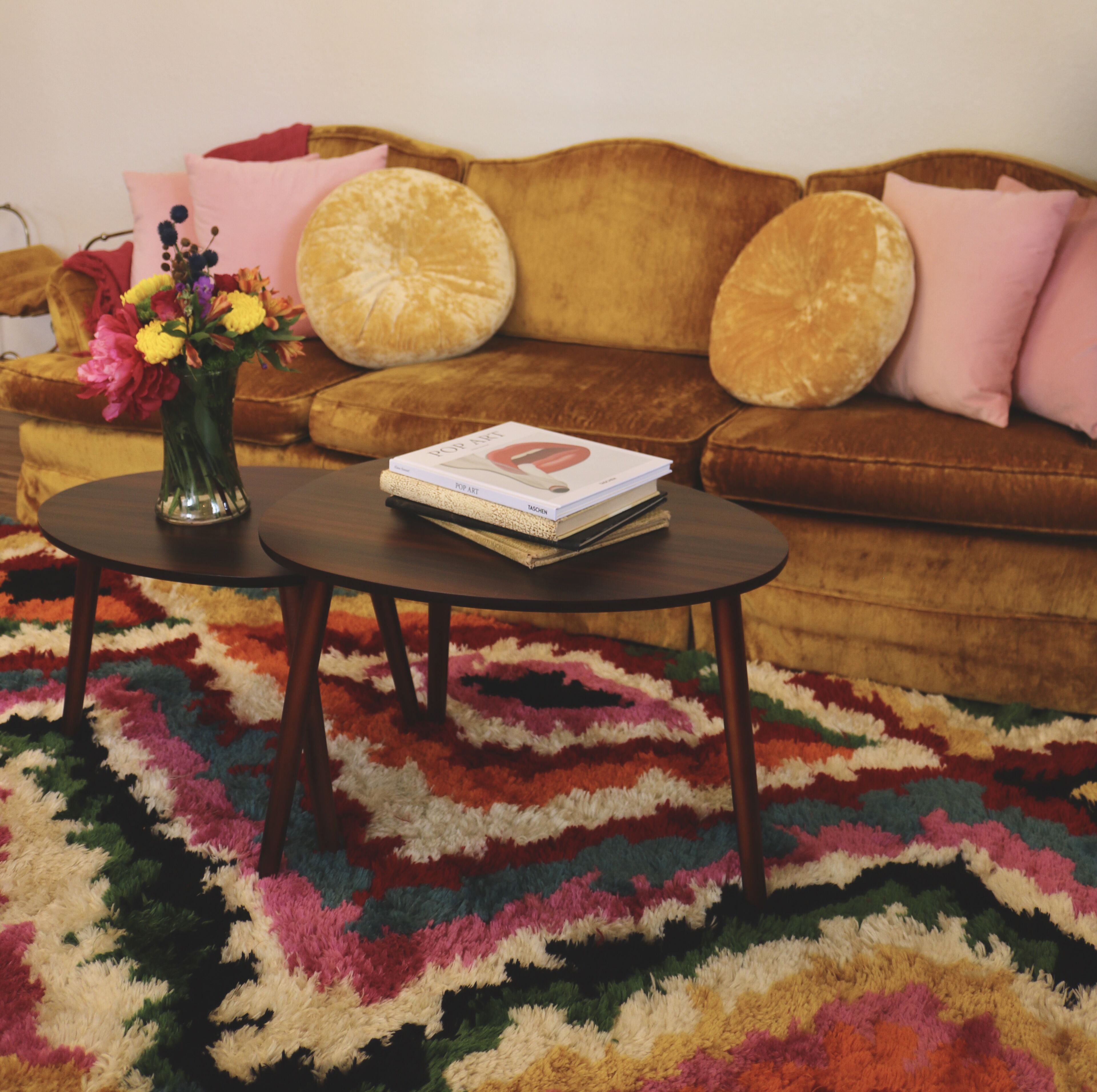 Un salon avec un canapé marron, deux tables rondes, un tapis coloré et un vase rempli de fleurs.