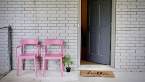 Deux chaises roses adossées à un mur de briques blanches, avec une porte et une petite plante en pot.