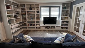Ett vardagsrum med en platt-TV, inbyggda hyllor med diverse dekorativa föremål, en blå soffa och en mönstrad matta.