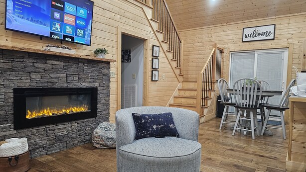A cozy living room with a stone fireplace, a flat-screen TV, a staircase, and a 'welcome' sign. - Sugar Bear Cabin 5 mins from State Park!  Fishing, w/boat parking, golf & more! (Burkesville)