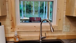 A kitchen with a double sink, wooden cabinets, a window with a view of trees, and a lace curtain. - Sugar Bear Cabin 5 mins from State Park!  Fishing, w/boat parking, golf & more! (Burkesville)