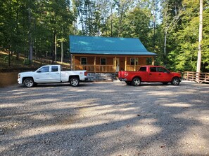 Une cabane avec une allée en gravier et deux camions garés, entourée d’arbres.