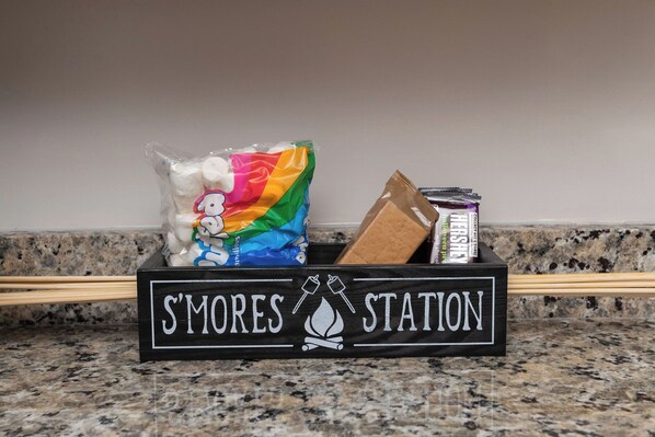 A kitchen counter with a black box labeled "S'MORES STATION" containing marshmallows, a Hershey's chocolate bar, and a bag of popcorn.