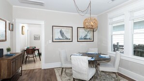 A dining area with a round table, white chairs, and a patterned rug. There are framed pictures on the wall and a hanging light fixture.