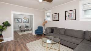 A modern living room with a gray sofa, a glass coffee table, a blue armchair, and a dining area with a chandelier.
