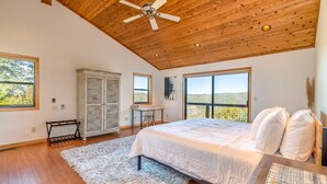 A bedroom with a wooden ceiling, a large bed, a desk, and a view of the outdoors.