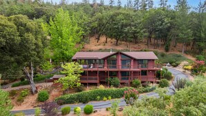 A two-story cabin with a large deck and multiple windows, surrounded by dense forest and a winding road.