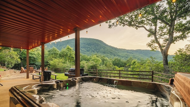 A hot tub area with a wooden deck, a covered seating area, and a view of a forested mountain.