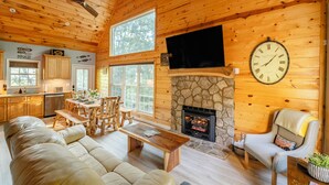 A log cabin interior with a stone fireplace, wooden furniture, a dining area, and a large clock on the wall.