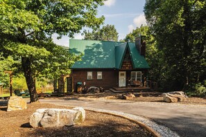 A rustic log cabin with a green roof, surrounded by trees and a gravel driveway.