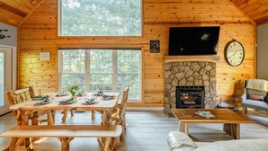 A wooden cabin interior with a dining table set, a stone fireplace, a television mounted on the wall, and a large window.